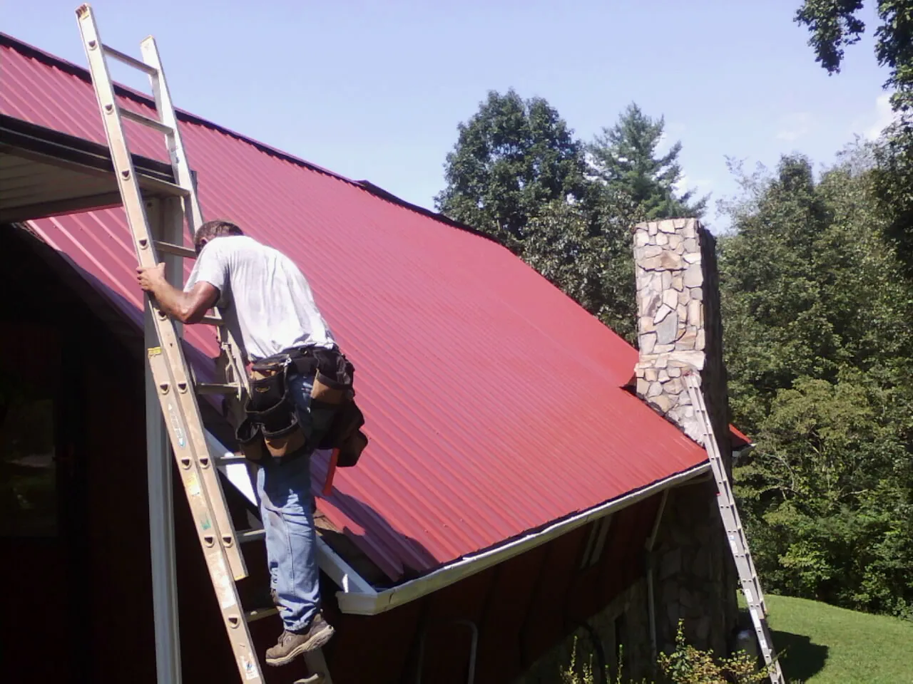 Roofer on a ladder beside a red metal roof and stone chimney — Soffit & Fascia Repair by Roofing Brothers in Fenton